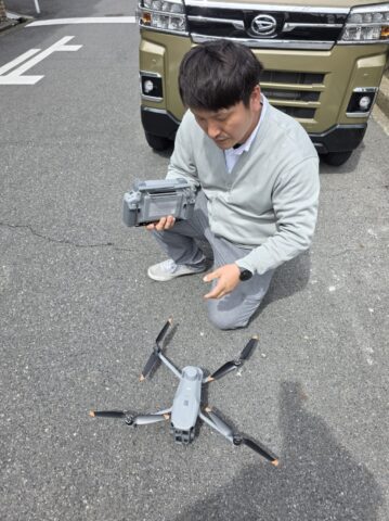 八尾市の青空のもと、サーモグラフィカメラを搭載した大型の産業用ドローンを飛行させている、二級建築士の職人